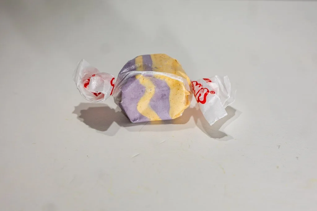 Blackberry Crumble flavored taffy. Clear wrapped with red lettering on the ends. purple and tan striped piece with specks in the tan sections.