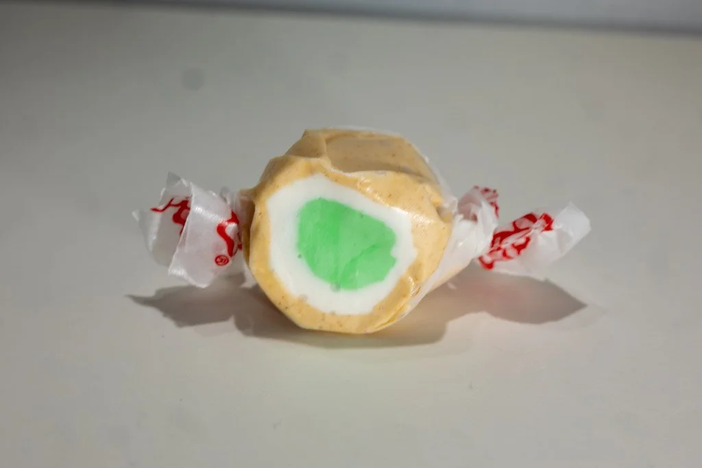 tan piece of taffy with specks of cinnamon, white, and a green apple flavored center. Wrapped in clear wrapper with red lettering on the end. Apple Pie flavored Taffy.