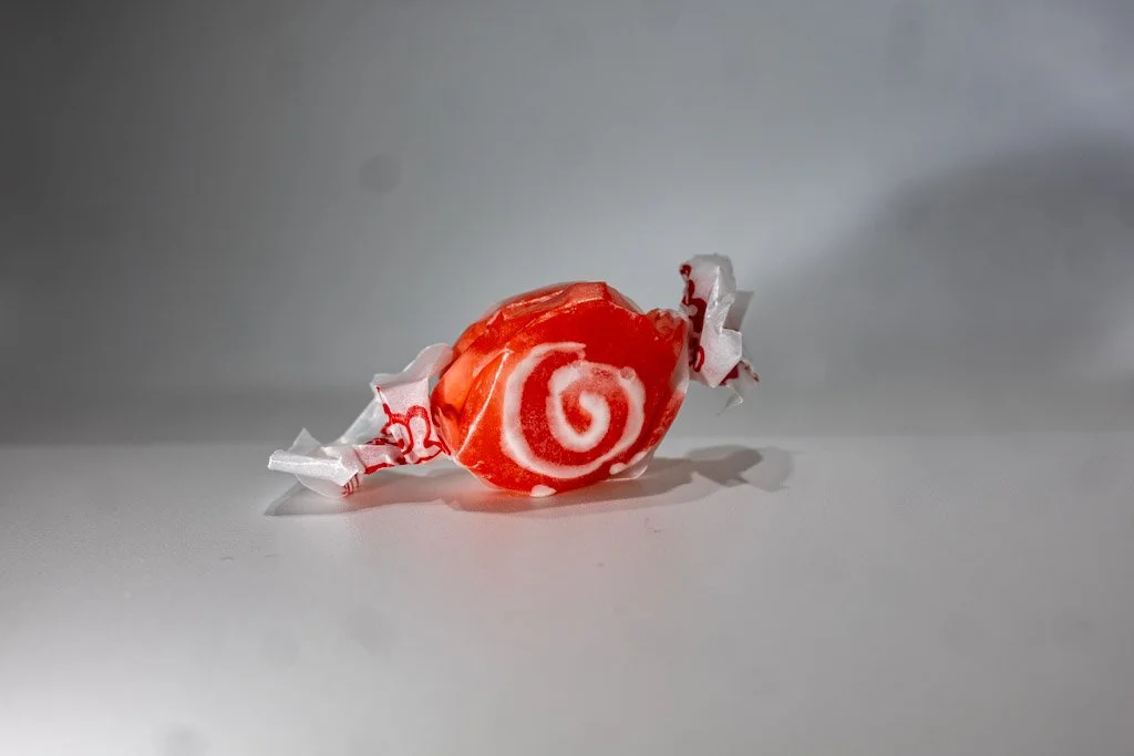 A wrapped piece of red licorice saltwater taffy with a vibrant red exterior and white swirl center, twist-wrapped in clear wax paper with red lettering on each end. A bold, chewy classic sold at Sea Bird Gifts & Candy in Westport, WA.
