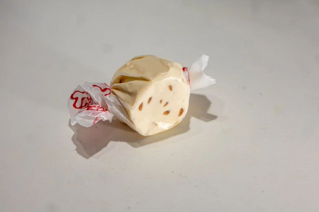 A wrapped piece of chocolate chip cookie saltwater taffy with a clear wax wrapper and red lettering on each twist end. The creamy beige candy has visible chocolate specks throughout