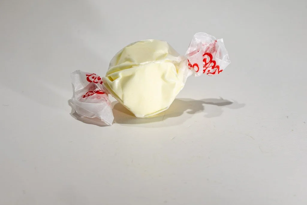A wrapped piece of egg nog saltwater taffy in a pale cream color, twist-wrapped in clear wax paper with red lettering on each end. A smooth, holiday-inspired flavor sold at Sea Bird Gifts & Candy in Westport, WA.