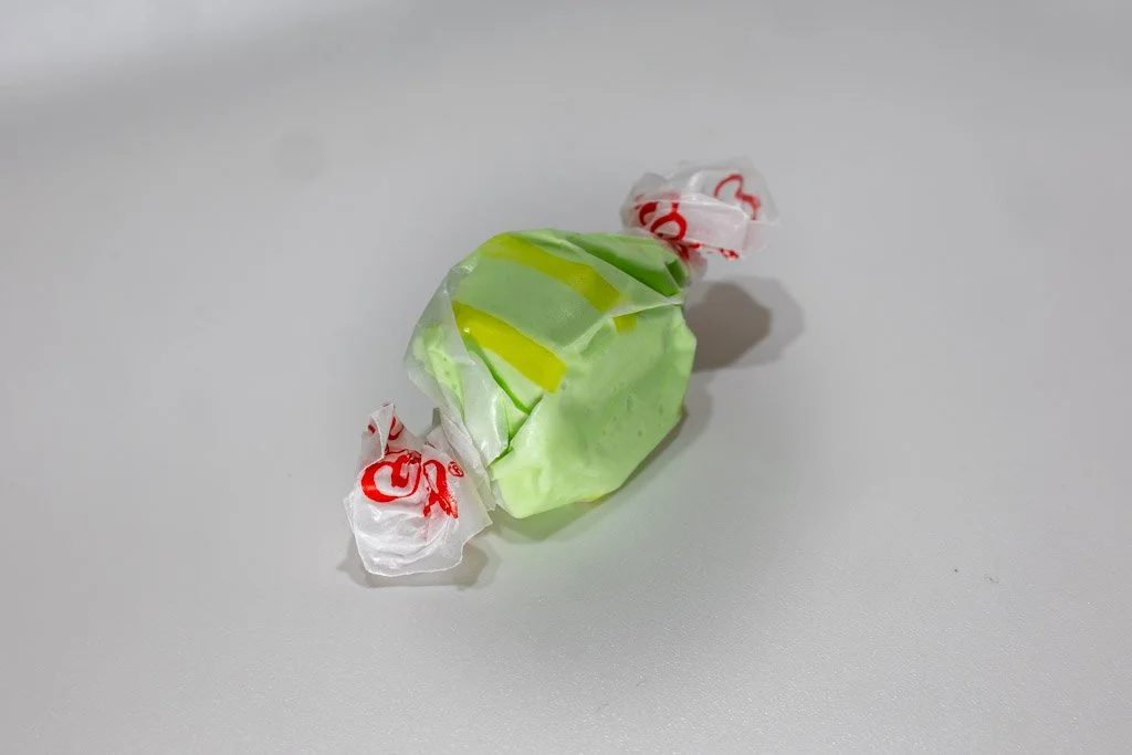 A wrapped piece of golden pear saltwater taffy in light green with vibrant yellow swirls, twist-wrapped in clear wax paper with red lettering on each end. A subtly sweet fruit-flavored taffy from Sea Bird Gifts & Candy in Westport, WA.