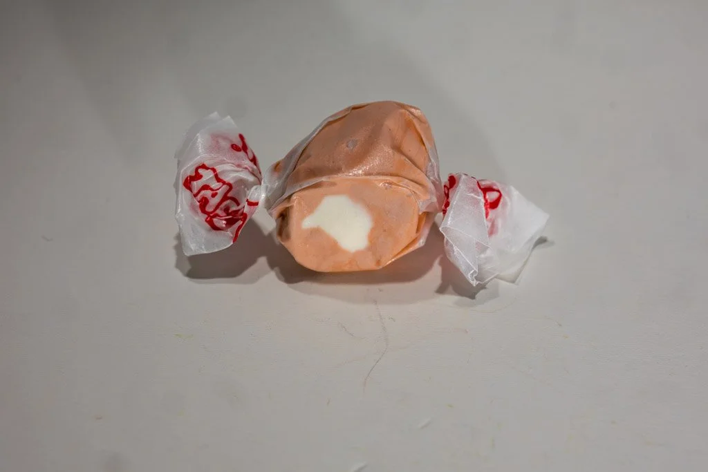 A wrapped piece of root beer float saltwater taffy with a warm brown exterior and a creamy white center, twist-wrapped in clear wax paper with red lettering on each end. A retro soda-inspired flavor from Sea Bird Gifts & Candy in Westport, WA.