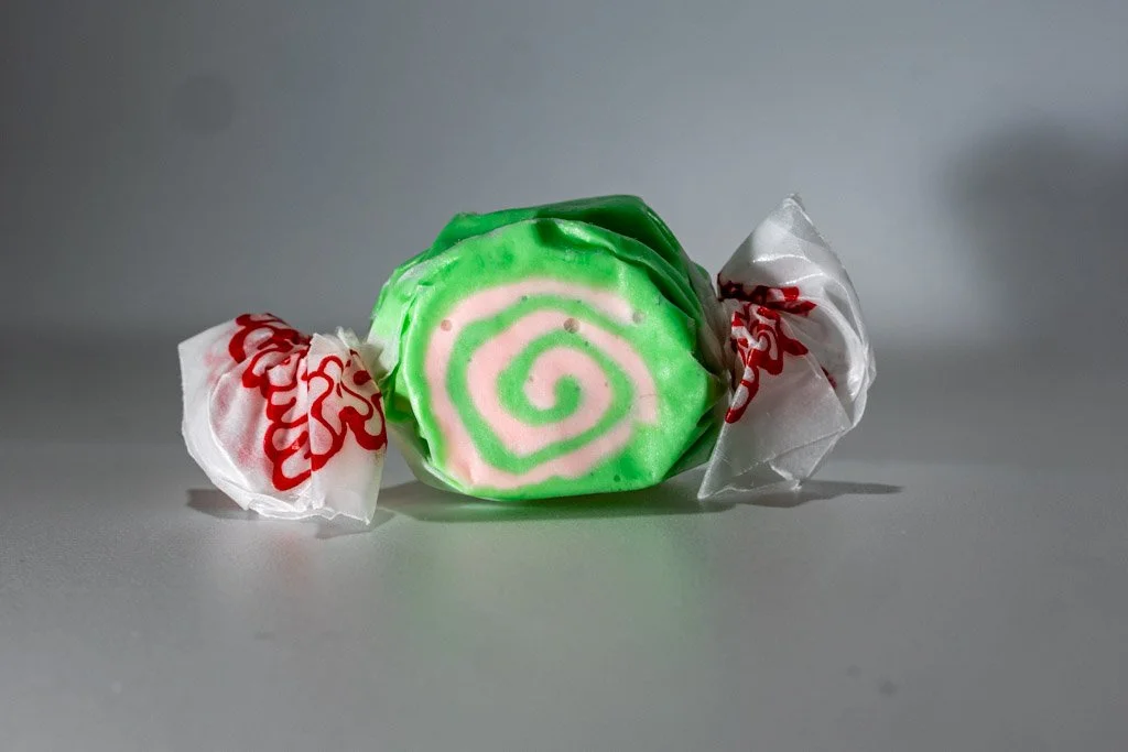 A wrapped piece of cherry limeade saltwater taffy with a vibrant pink and green spiral pattern, twist-wrapped in clear wax paper with red lettering on each end. A colorful, citrus-cherry flavored treat from Sea Bird Gifts & Candy in Westport, WA
