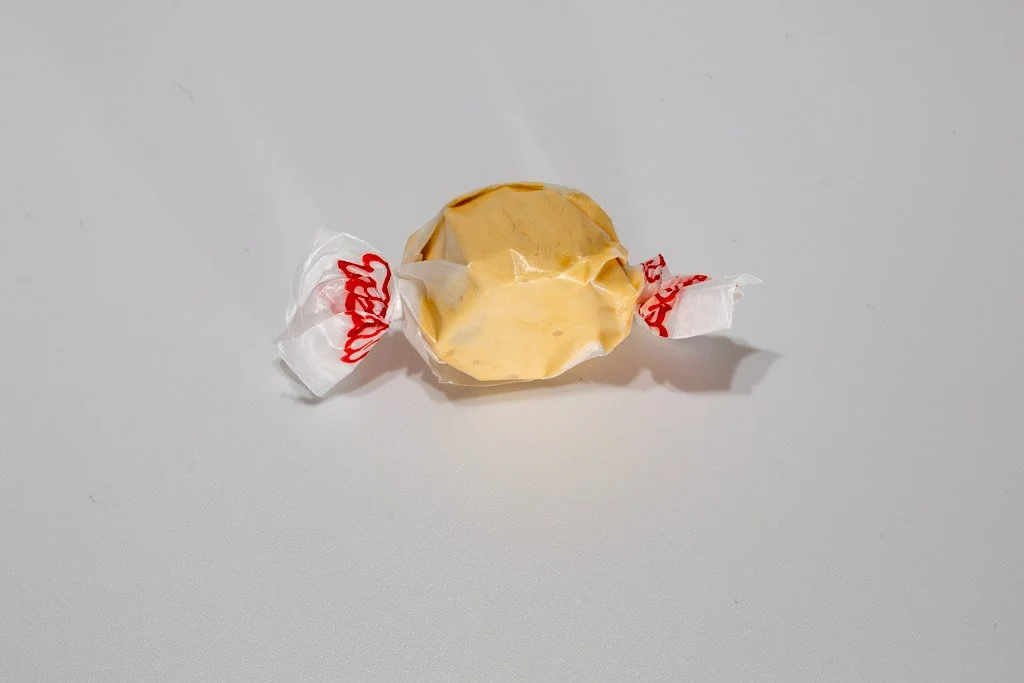 A soft, golden-yellow piece of taffy twist-wrapped in translucent wax paper with red print. Its buttery hue hints at the warm, caramelized flavor of classic butterscotch