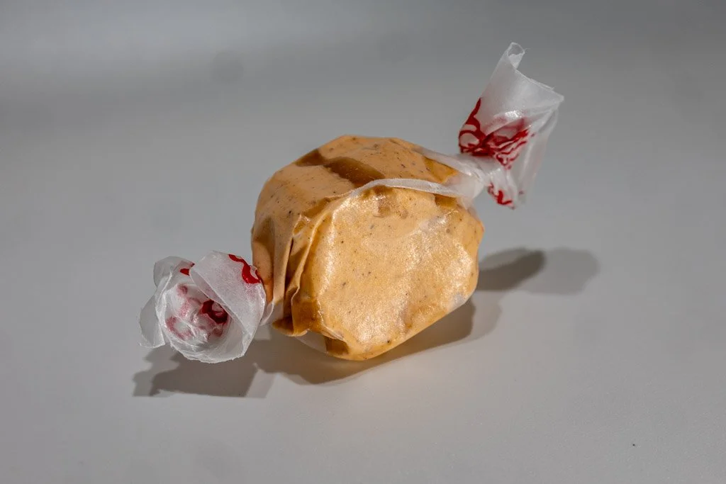 A wrapped piece of pumpkin pie saltwater taffy in a warm orange color with subtle cinnamon-brown specks, twist-wrapped in clear wax paper with red lettering on each end. A cozy, spiced fall favorite from Sea Bird Gifts & Candy in Westport, WA.