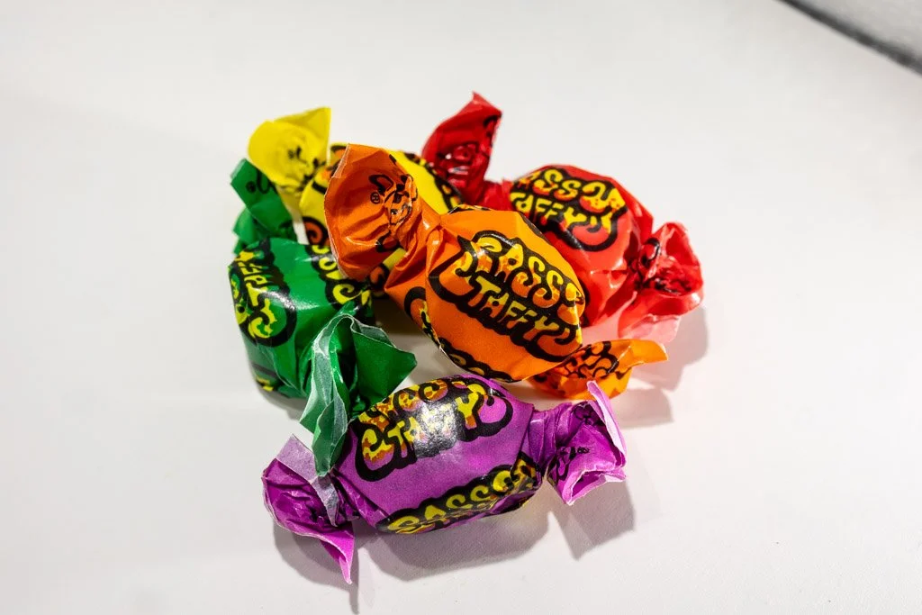 A colorful assortment of Sassy Taffy pieces in bold wrappers — orange, red, yellow, green, and purple — each piece twist-wrapped and labeled “Sassy Taffy” in a playful, sour-themed font