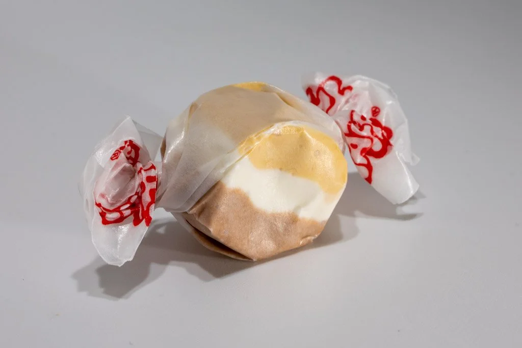 A piece of salt water taffy with distinct layers of brown, white, and tan, wrapped in wax paper printed with red text.