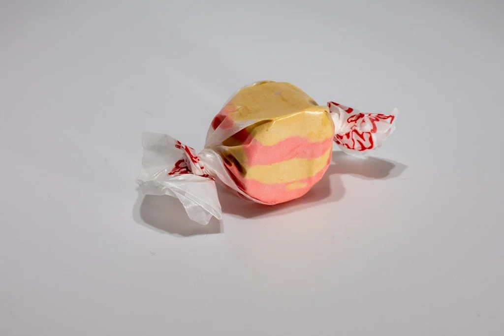A piece of saltwater taffy with swirls of warm golden yellow and smoky pink, twist-wrapped in clear wax paper with red print. The candy mimics the look of maple and bacon stripes — a novelty flavor from Sea Bird Gifts & Candy in Westport, WA.