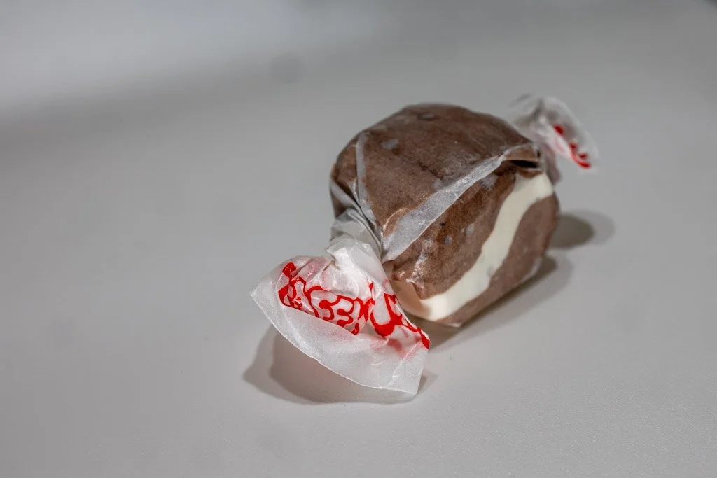A wrapped piece of cookies & cream saltwater taffy with dark brown and white swirled stripes, twist-wrapped in clear wax paper with red lettering on each end. A dessert-inspired candy from Sea Bird Gifts & Candy in Westport, WA.