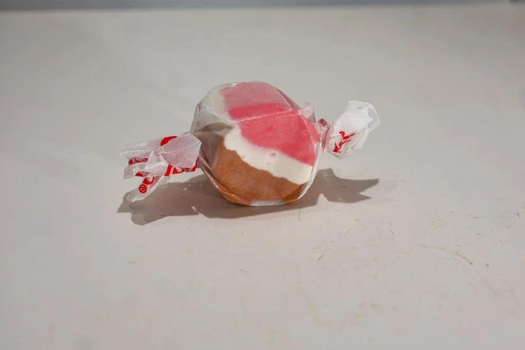 A wrapped piece of Neapolitan saltwater taffy with visible pink, white, and brown stripes, representing strawberry, vanilla, and chocolate. Wrapped in a clear wax wrapper with red lettering on each end.