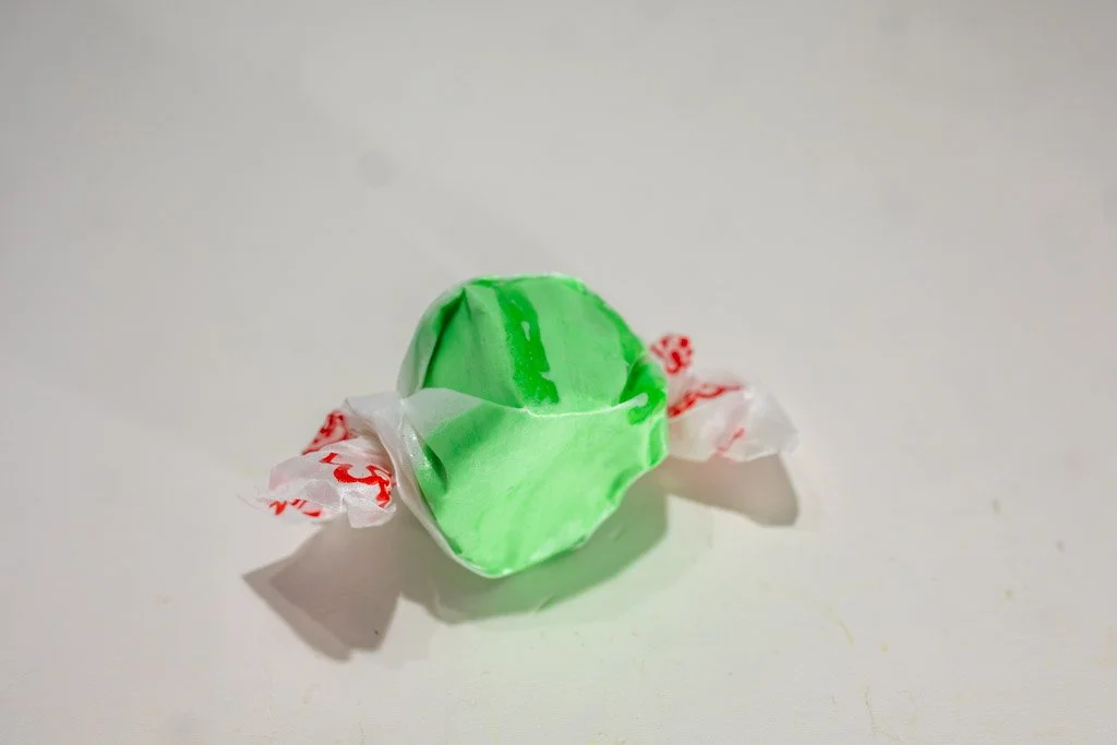 A wrapped piece of green apple saltwater taffy with light and dark green swirls, twist-wrapped in a clear wax wrapper with red lettering on each end. A tart and tangy favorite from Sea Bird Gifts & Candy in Westport, WA.