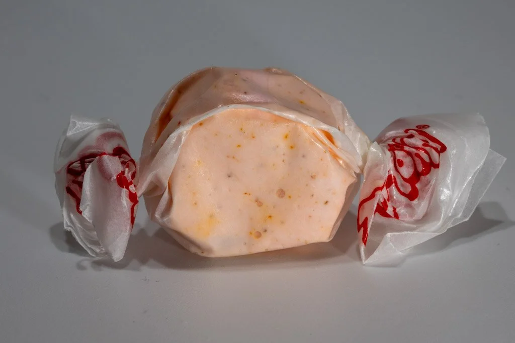 A round salt water taffy wrapped in clear wax paper, light orange in color with scattered red-orange specks throughout.
