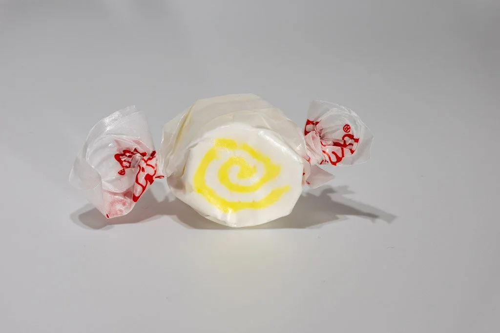 A white salt water taffy with a bright yellow swirl in the center, wrapped in clear wax paper with red print. The colors suggest a tropical, pineapple-coconut flavor