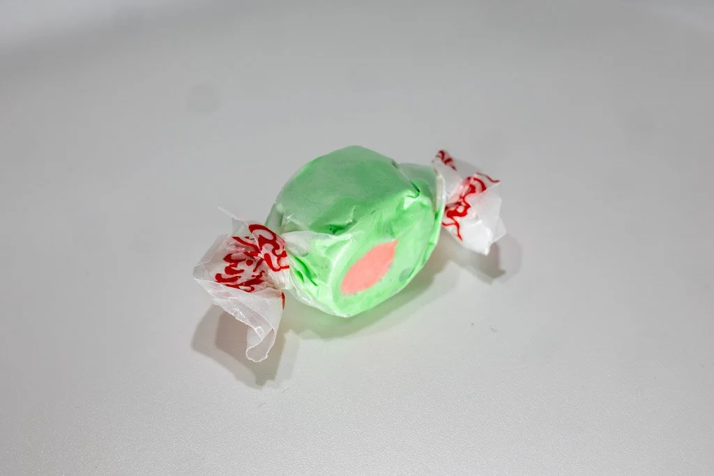 A wrapped piece of watermelon saltwater taffy with a bright green exterior and a soft pink center, twist-wrapped in clear wax paper with red lettering on each end. A juicy, two-tone fruit-inspired candy from Sea Bird Gifts & Candy in Westport, WA.