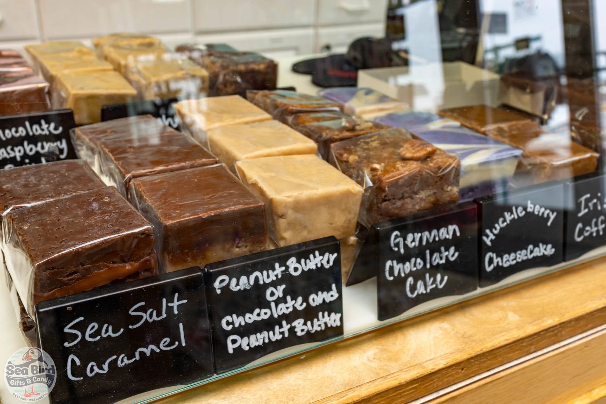 Assorted homemade chocolate and caramel fudge displayed behind glass at a candy shop, with black handwritten labels for different flavors including sea salt caramel, peanut butter, german chocolate, huckleberry cheesecake, and Irish coffee.