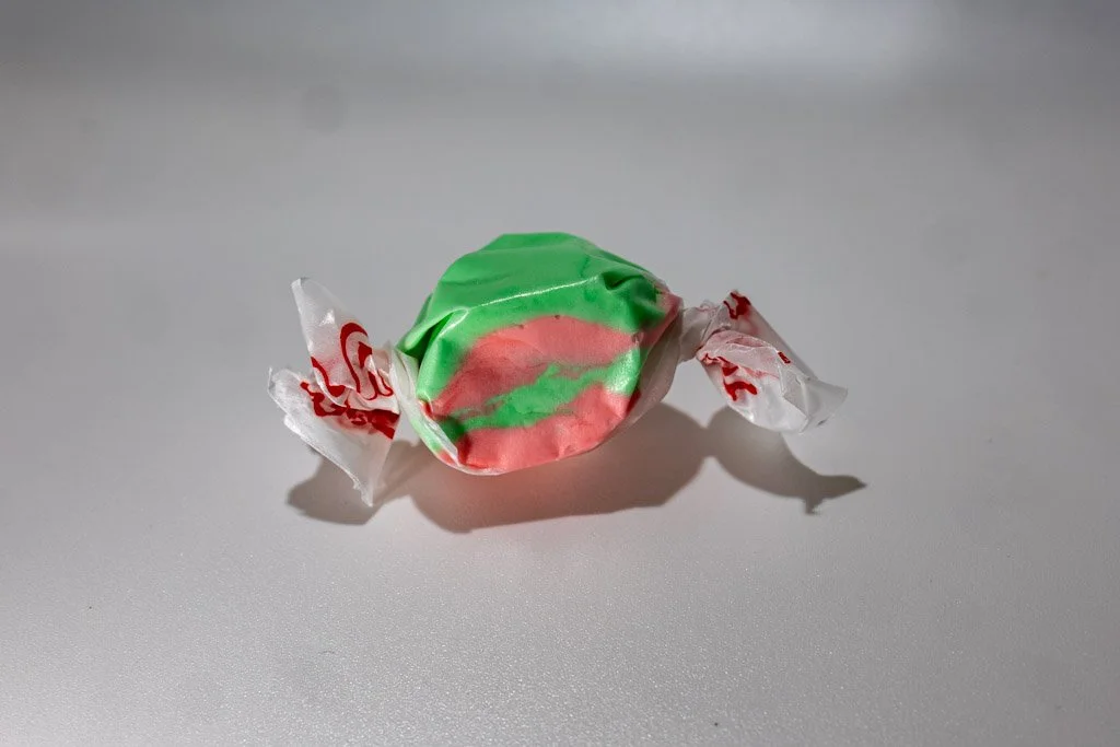 A wrapped piece of kiwi strawberry saltwater taffy with swirled pink and green coloring, twist-wrapped in clear wax paper with red lettering on each end. A bright, fruity combo candy from Sea Bird Gifts & Candy in Westport, WA.