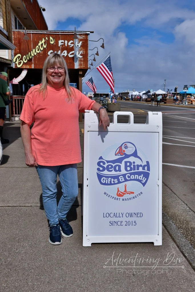 Paula Bird, owner of Sea Bird Gifts & Candy in Westport, WA, standing beside the locally owned candy and gift shop sign near the Westport marina