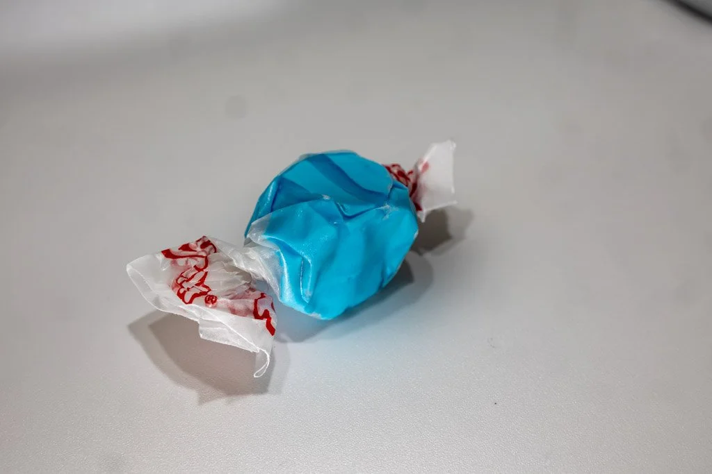 A wrapped piece of blue raspberry saltwater taffy in a vibrant solid blue color, twist-wrapped in clear wax paper with red lettering on each end. A bright, fruity favorite sold at Sea Bird Gifts & Candy in Westport, WA