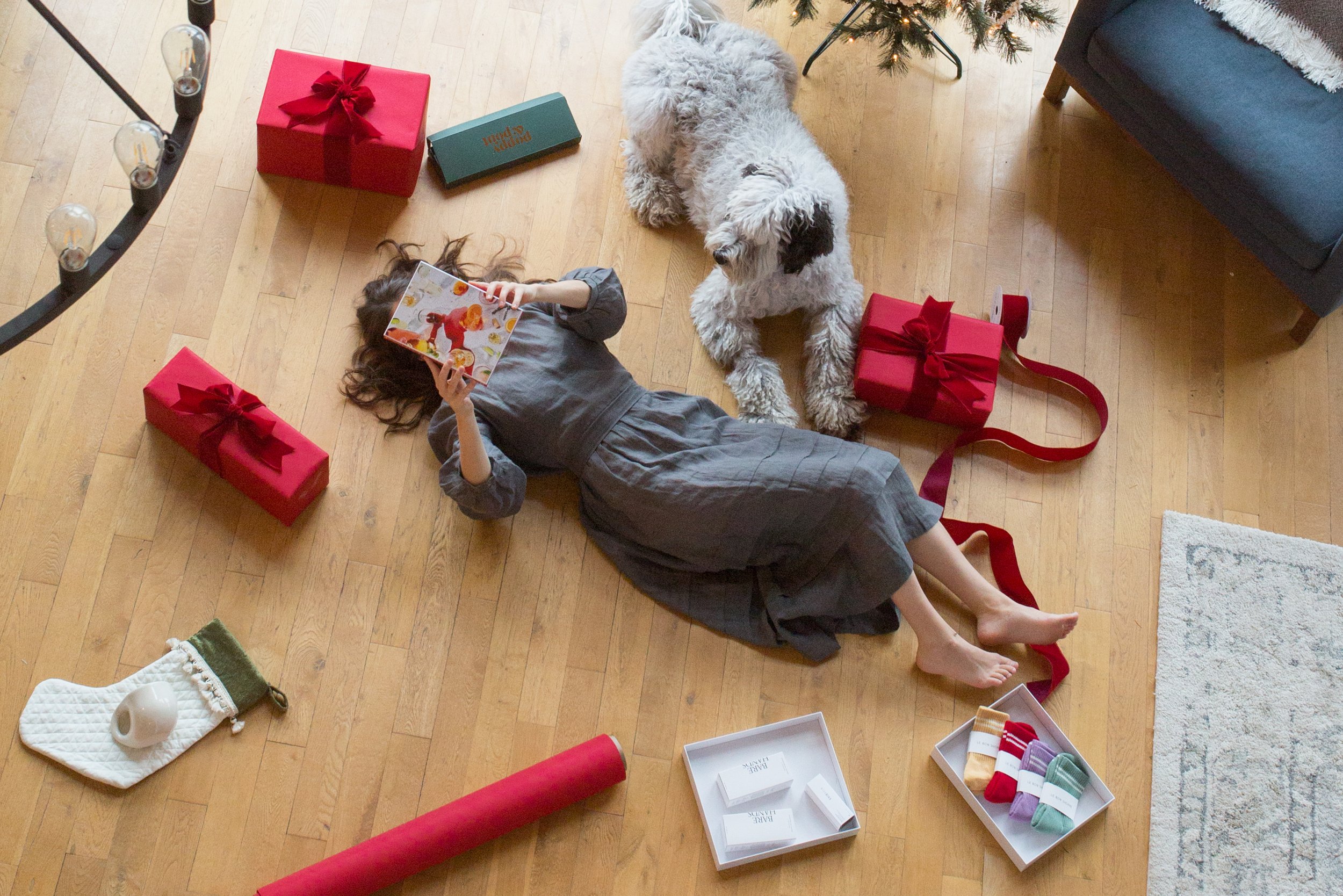 Holiday gift guide featuring a woman relaxing beside her dog, surrounded by wrapped presents and curated holiday gifts.