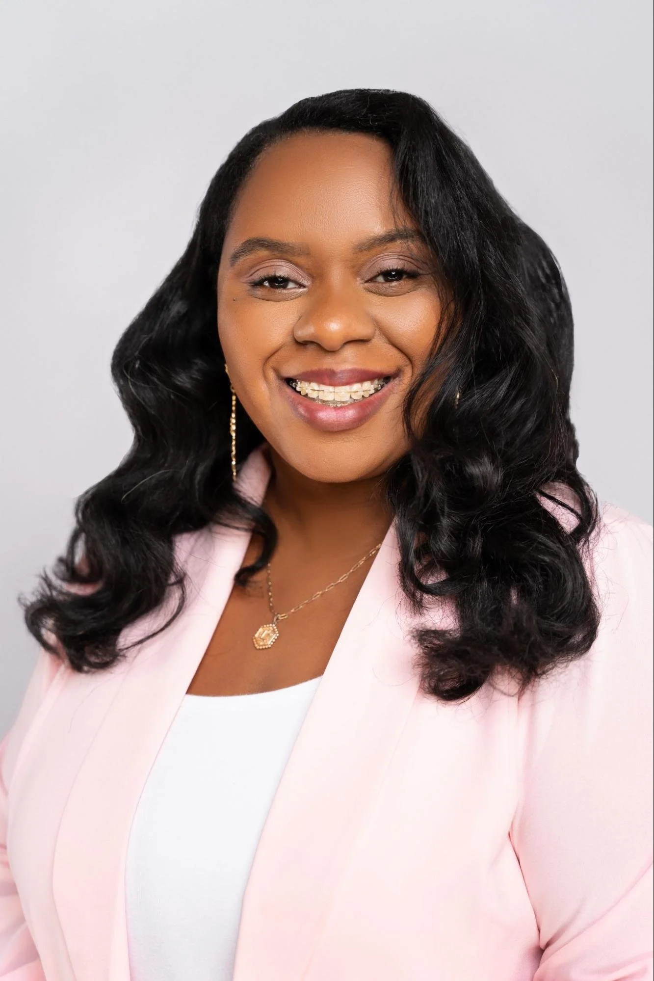 A woman smiling, wearing a pink blazer, white top, gold jewelry, and with long black wavy hair on a plain white background.