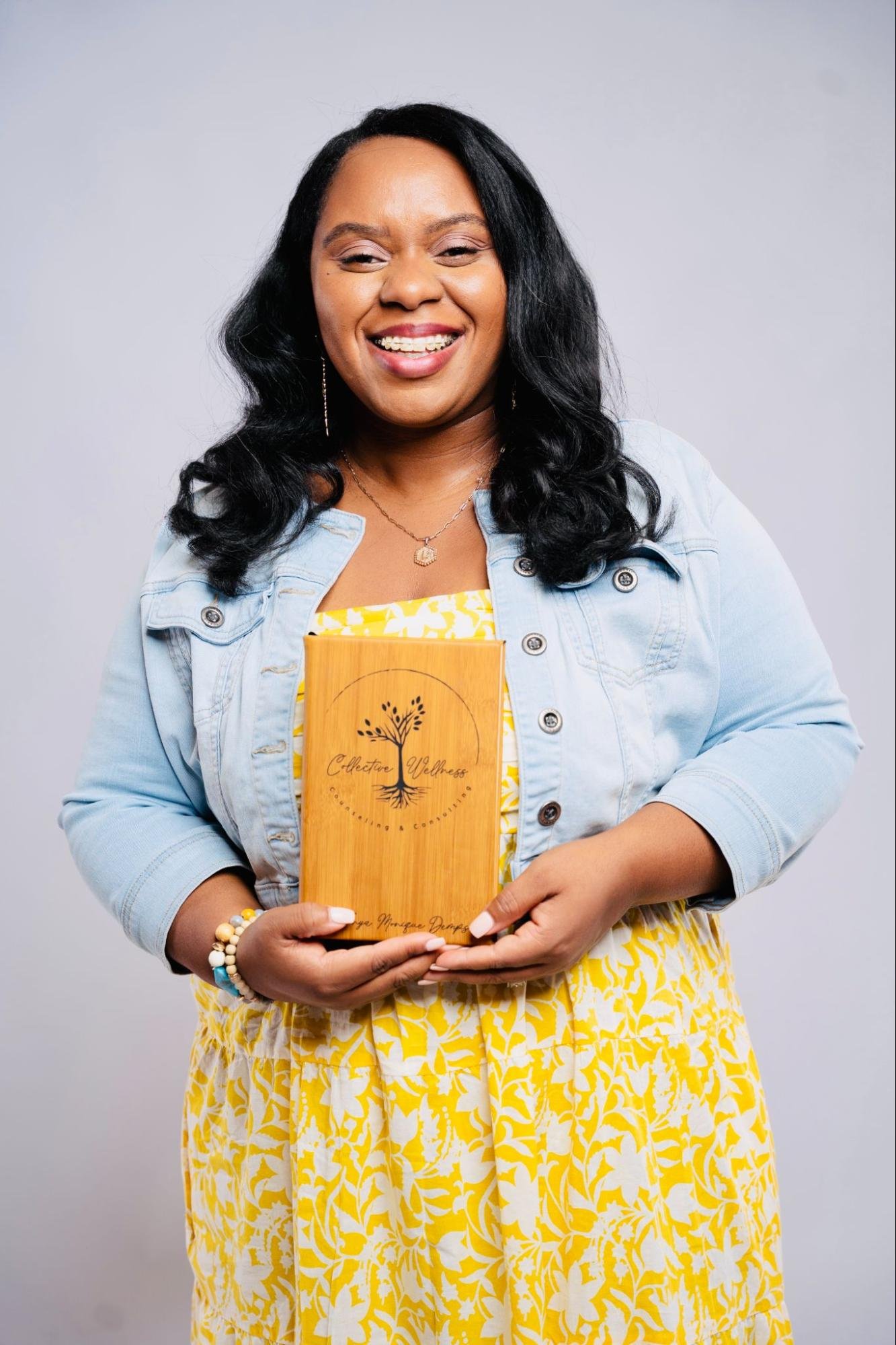 A woman with long black hair, wearing a yellow and white dress and a light denim jacket, is smiling and holding a wooden book titled 'Collective Wellness' with her hands. She is standing against a plain grey background.