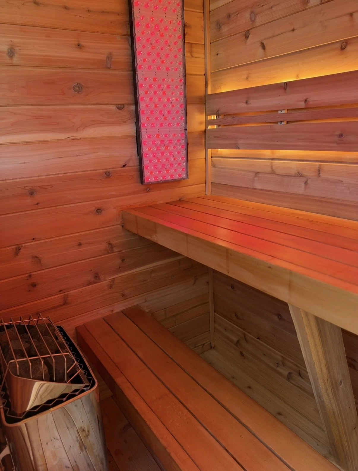 Wooden sauna interior with tiered benches, a sauna heater, and red infrared light panel