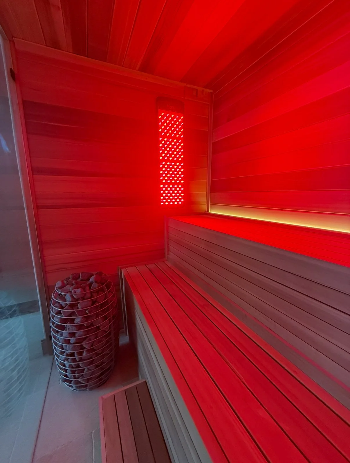 Inside a sauna with wooden benches and walls, red lighting, and a basket of stones in the corner.