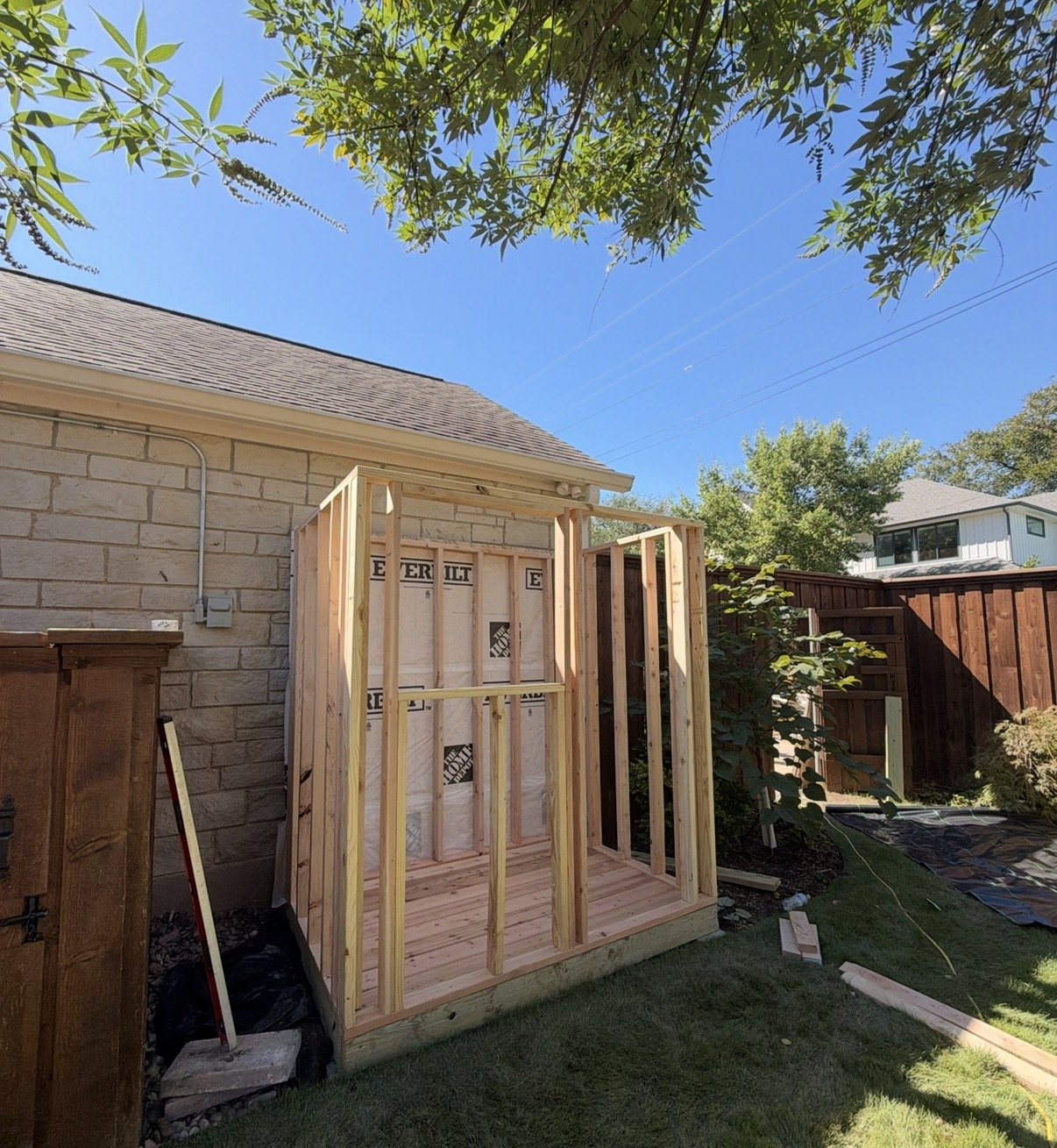 Partially framed backyard shed addition under construction beside a stone house, with exposed lumber, insulation, lawn, trees, and fencing.
