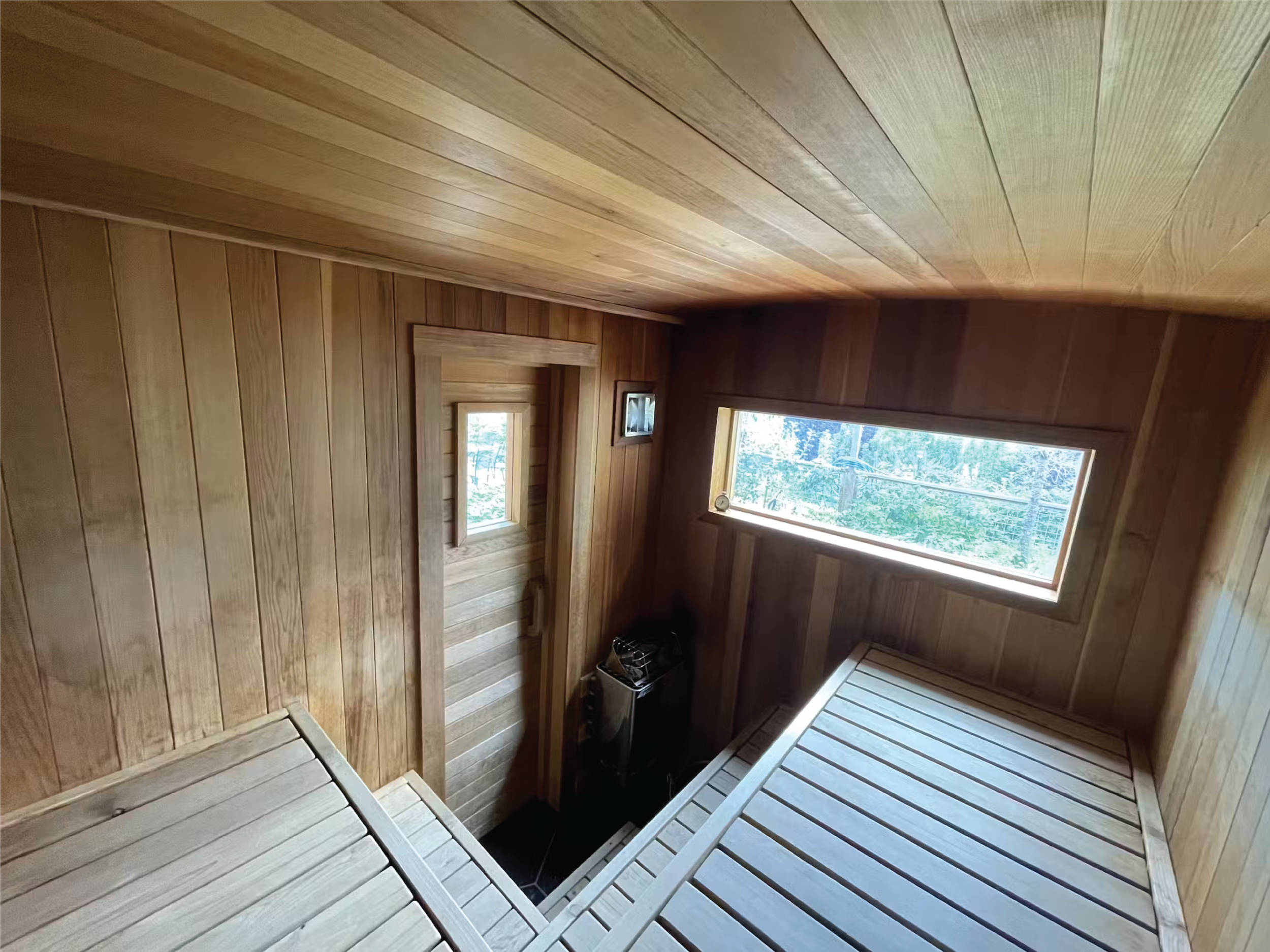Wood-paneled sauna interior with tiered benches, a compact heater, and windows overlooking trees.