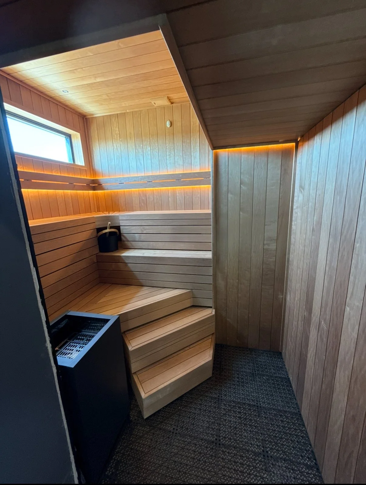 Wood-paneled sauna room with benches, a sauna bucket, and a heater, illuminated by soft LED lighting near a small window.