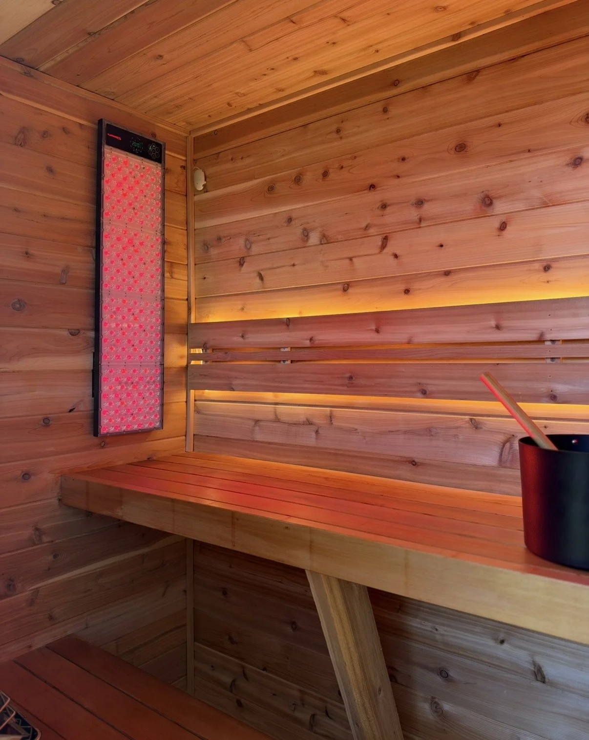 Wood-paneled sauna interior with a glowing red light therapy panel, wooden bench, and black sauna bucket.