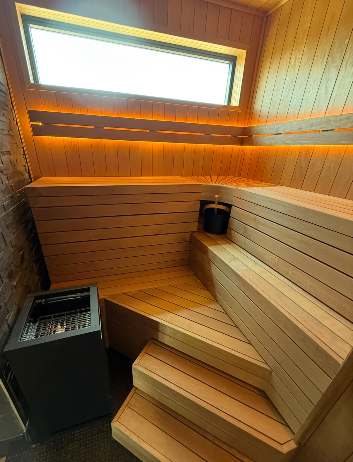 Wood-paneled home sauna with tiered benches, warm LED lighting, window, and black sauna heater.