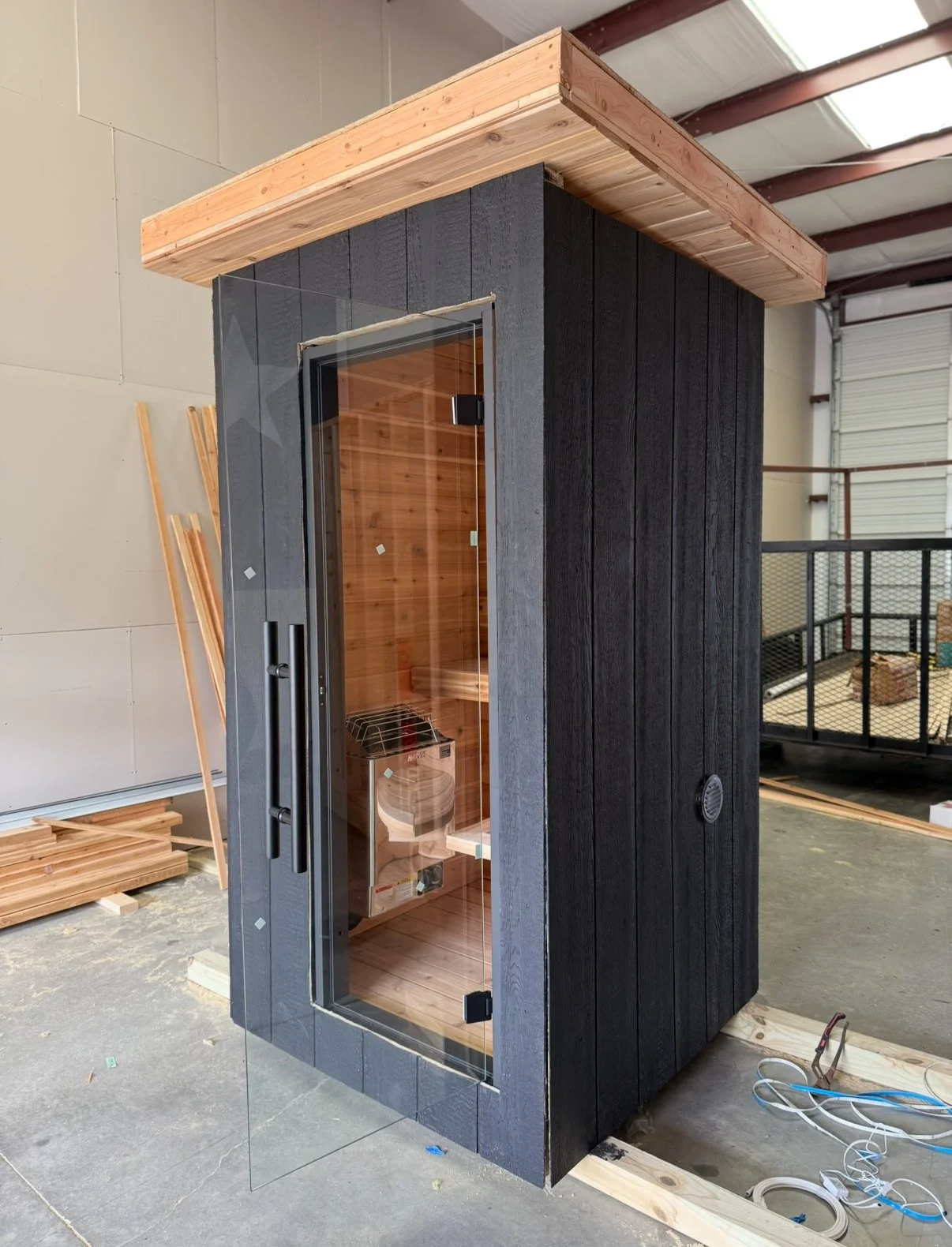 Small sauna with black exterior and glass door, located inside a warehouse under construction.