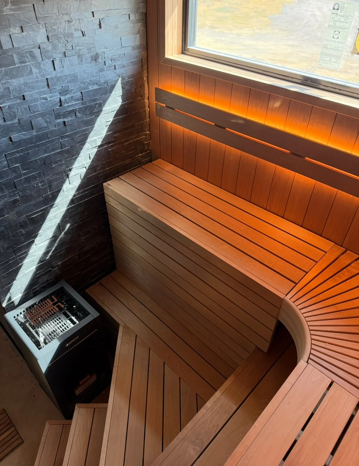 Interior of a modern sauna with wooden benches, a stone accent wall, a heater, and a window with natural light streaming in.