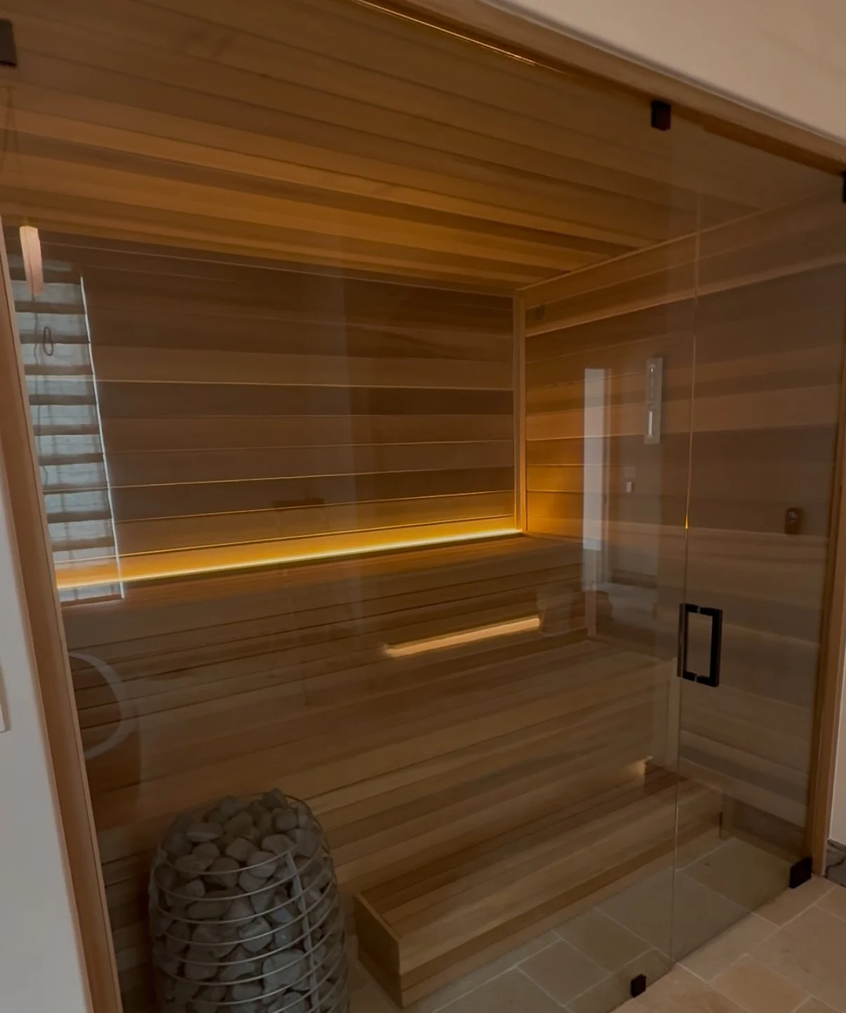 A modern indoor sauna with wooden interior, glass door, and a corner with stones inside a wire basket.