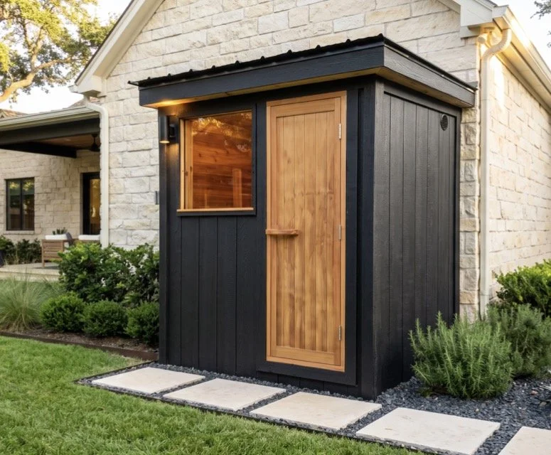 Small black-and-wood backyard sauna shed beside a light stone house, surrounded by landscaping.