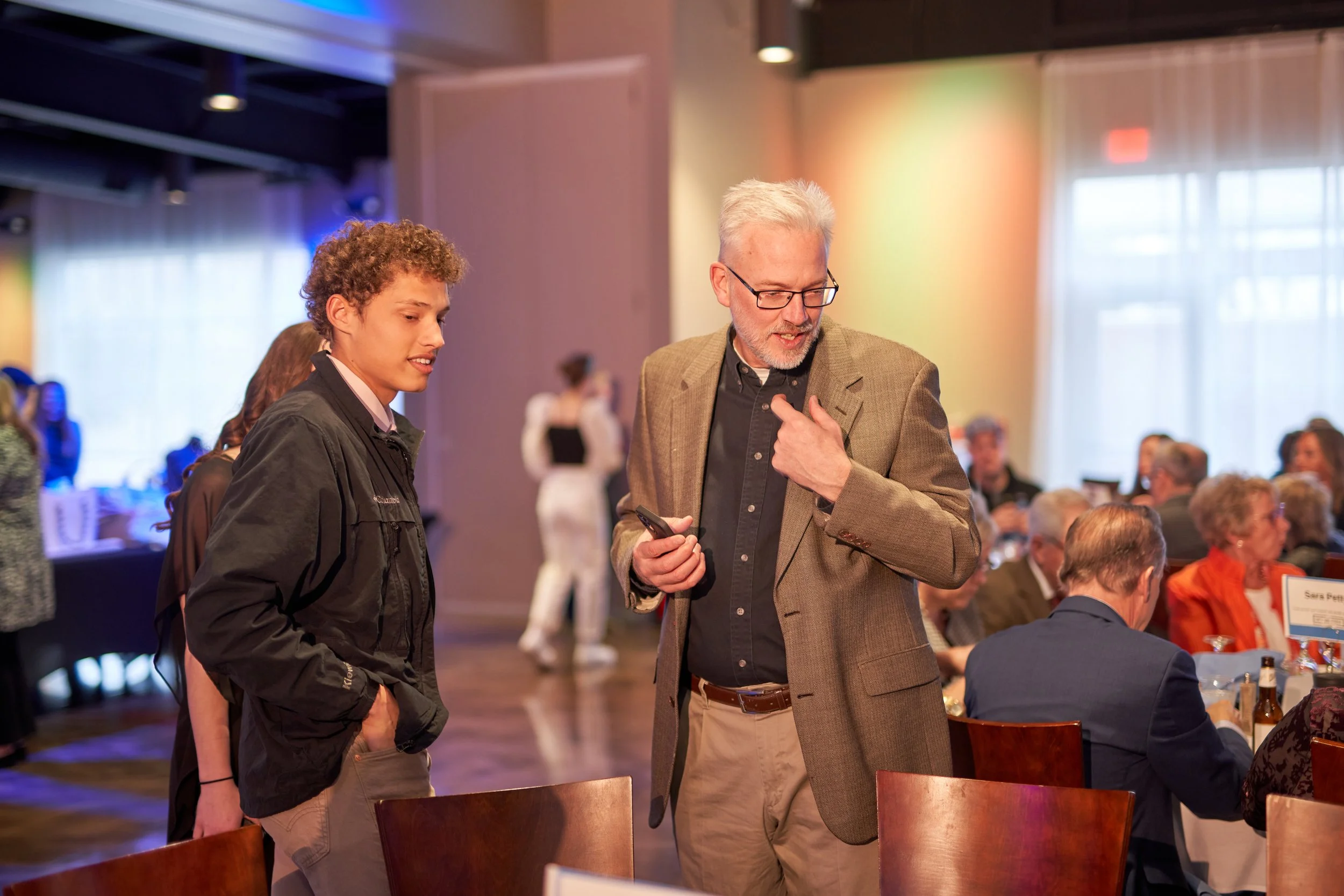 A man in a suit and a young man in a jacket having a conversation at a formal event. Tables with seated guests are visible in the background.
