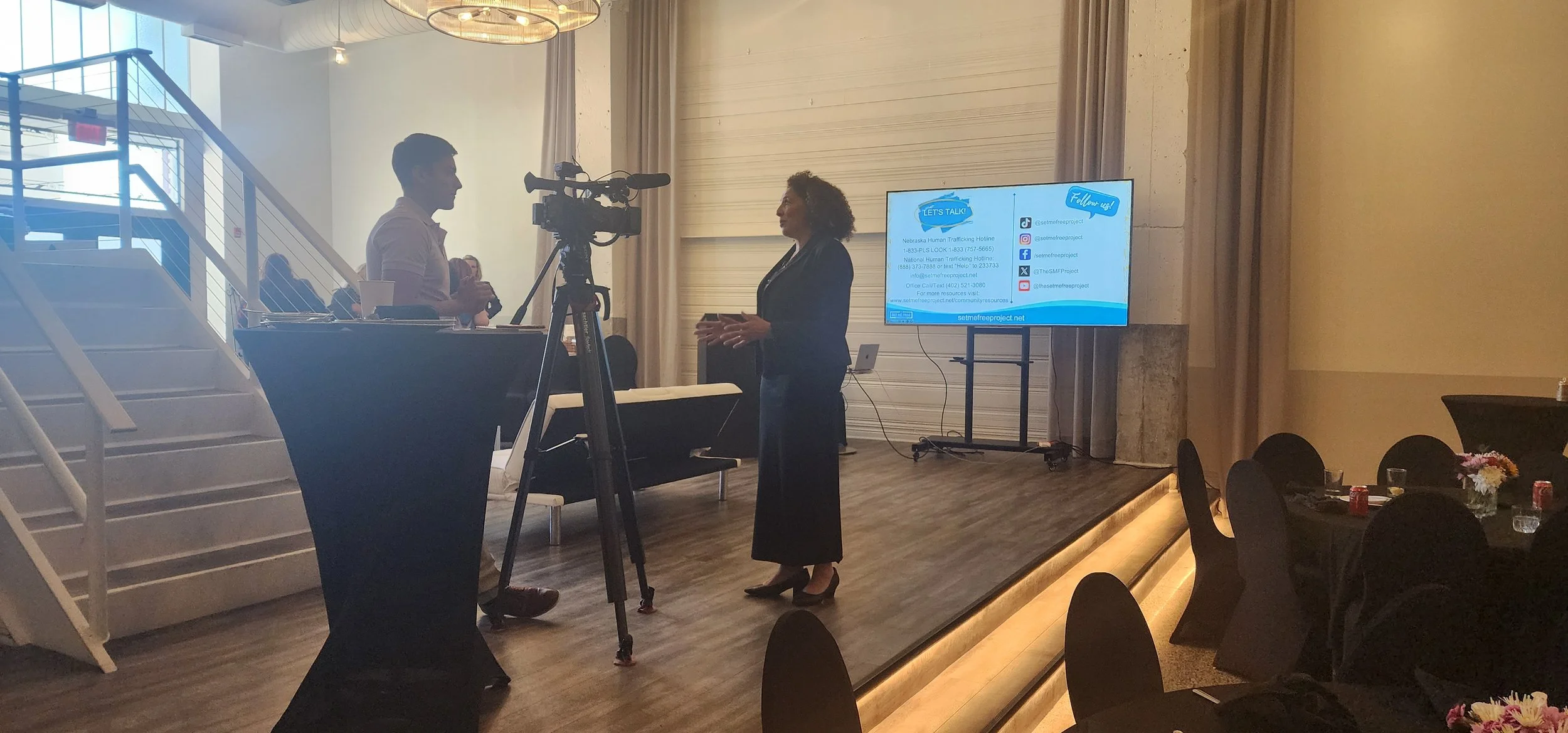 Two people talking in a conference room with a camera on a tripod, TV screen displaying contact information for "Set Me Free Project," tables with chairs and flower decorations, and stairs nearby.
