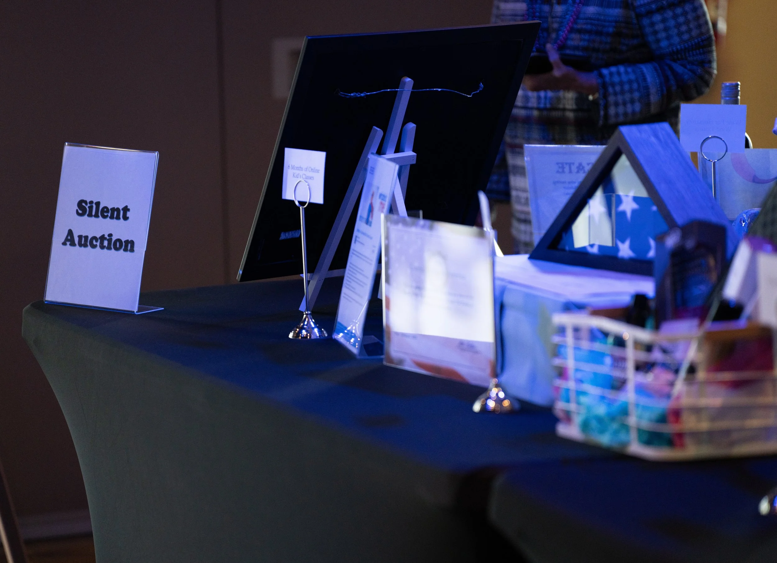 Silent auction table with various items displayed, including a sign with 'Silent Auction', framed photographs, and a basket with colorful items.