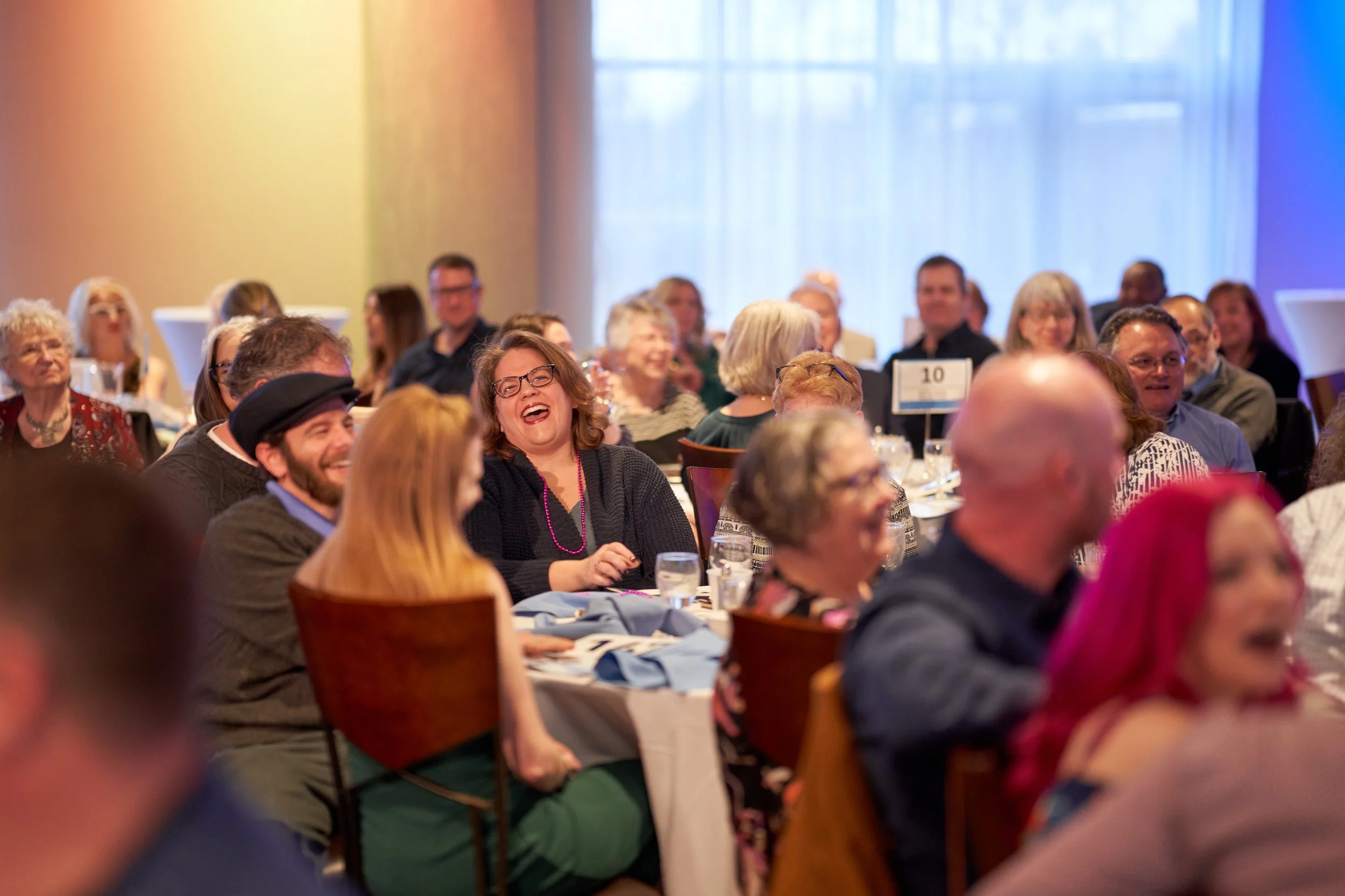 Group of people seated at round tables in a banquet setting, laughing and conversing.