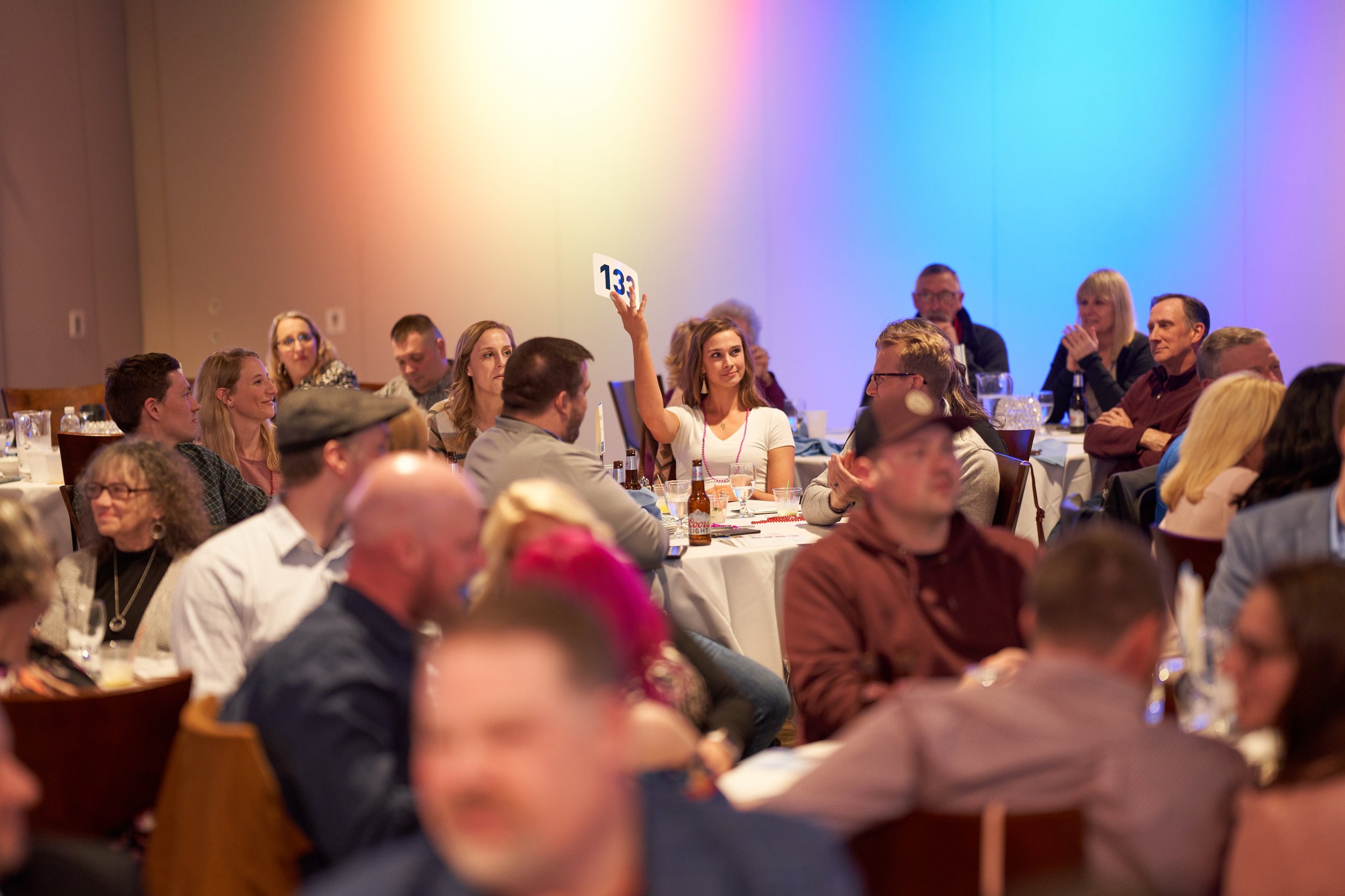 People seated at tables at an indoor event, one person holding up a bidding paddle with the number 134 in a lively auction setting.