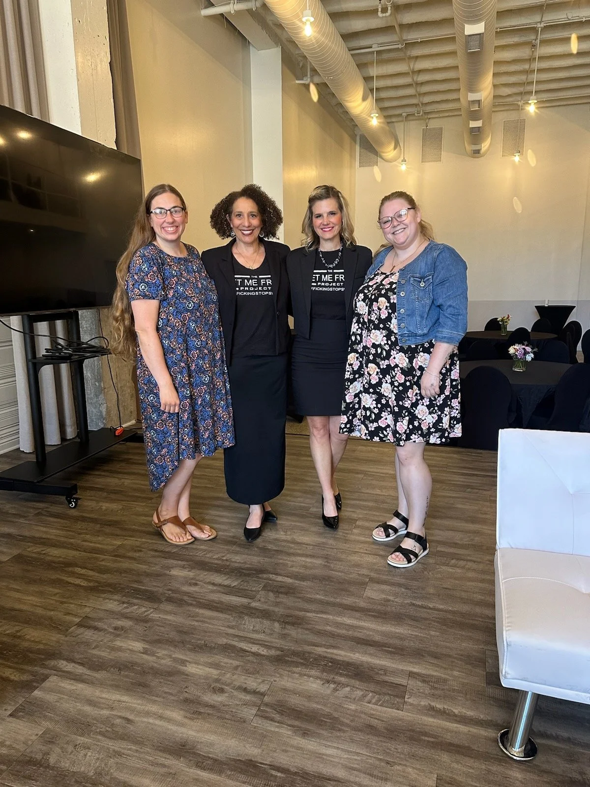 Four women standing together indoors, smiling at the camera. Two women are wearing floral dresses, the other two are in matching black outfits with printed T-shirts. The room has a wooden floor, a large TV screen, and visible ceiling ducts.