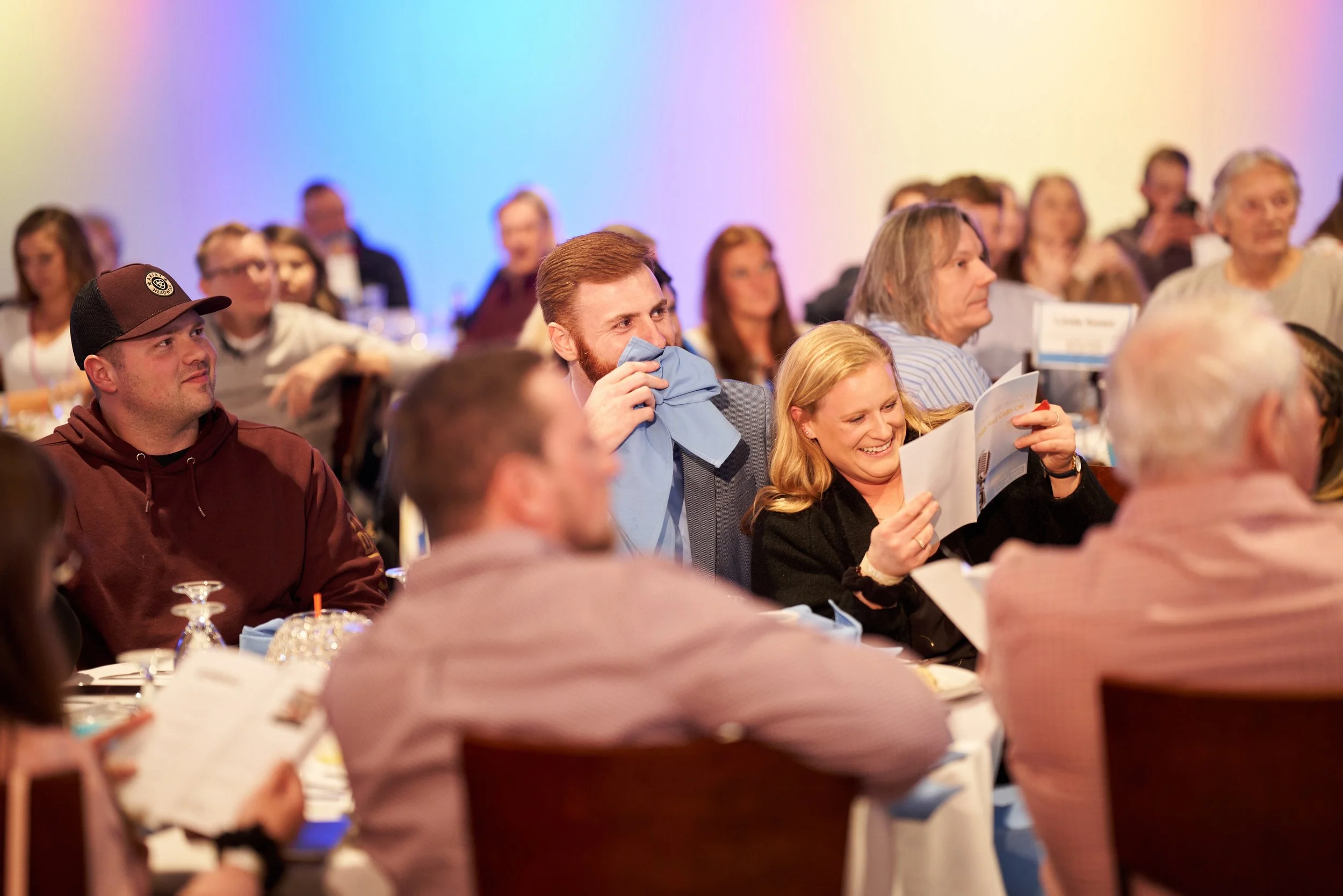 People attending a social event seated at tables, some holding papers, colorful lighting in the background.
