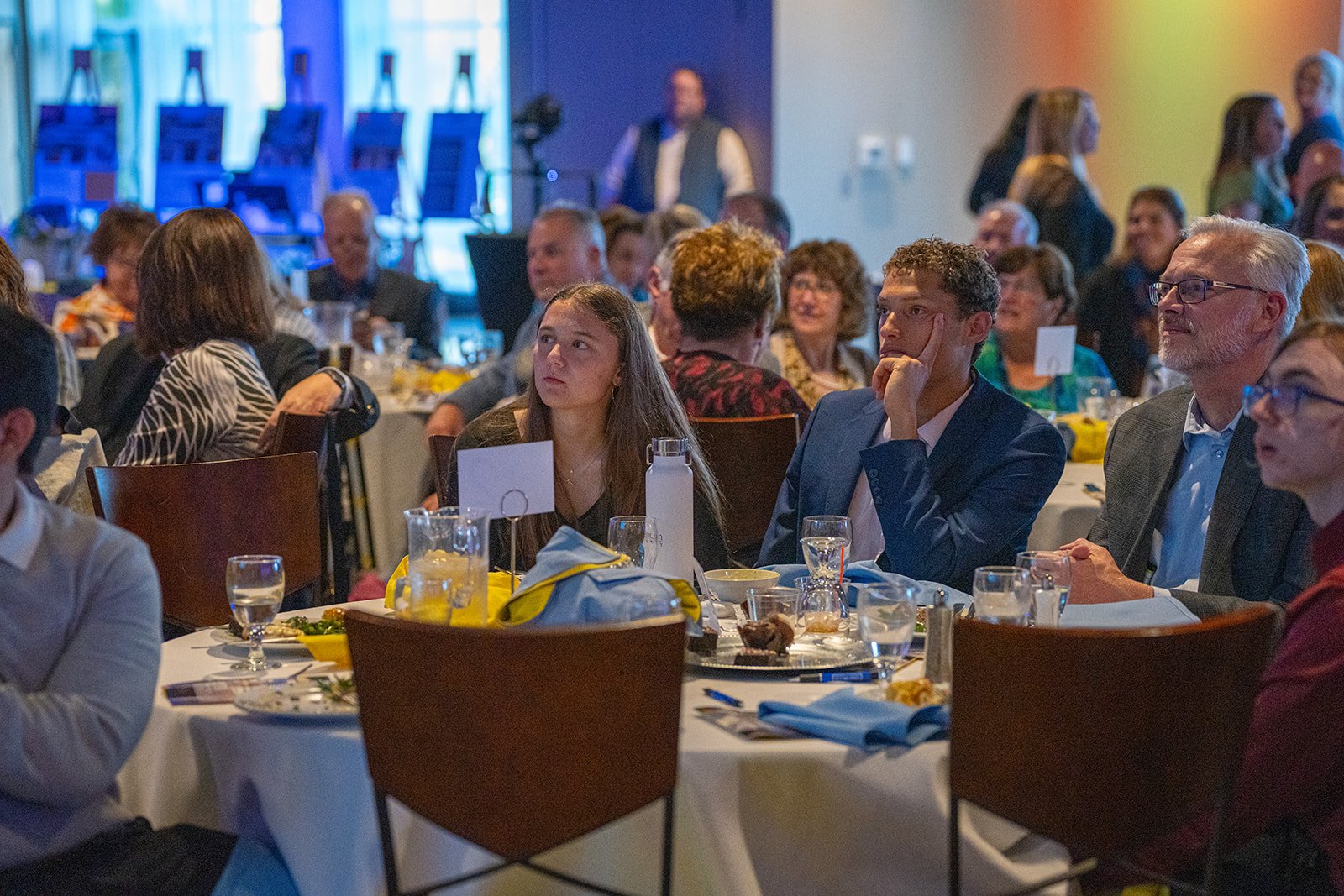 People attended The Set Me Free Project's fundraiser at round tables covered with white tablecloths. The attendees, including a young woman and a man in a blue suit, appear focused on a presentation or speaker. 