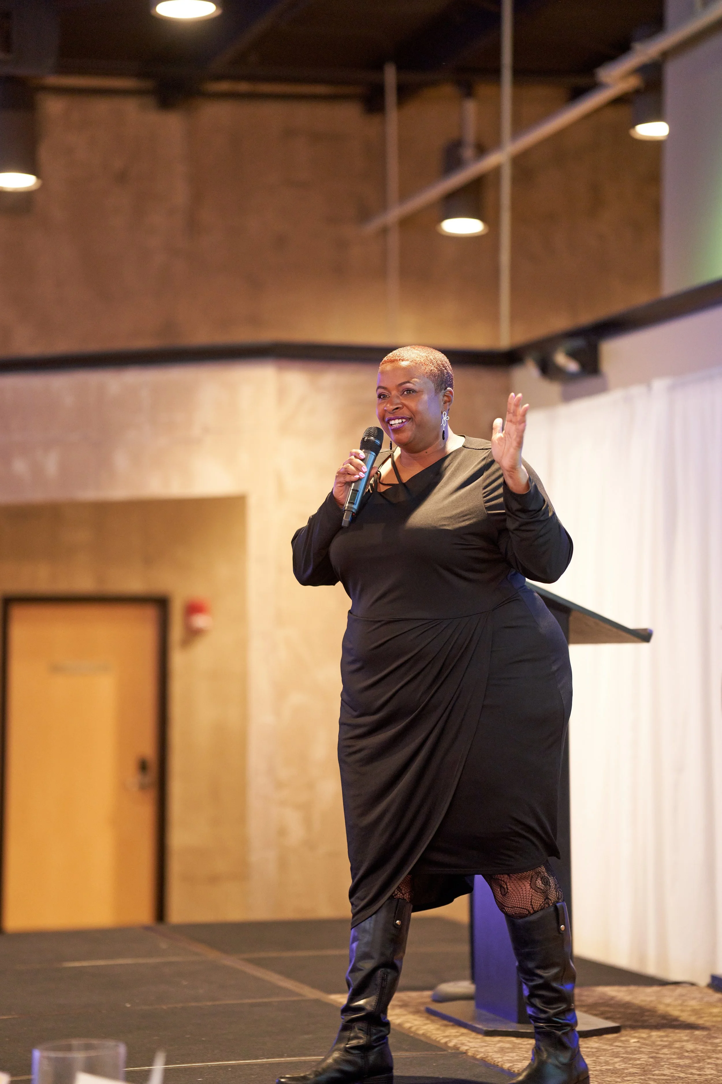 Person speaking on stage with microphone in hand, wearing a black dress and boots, in a well-lit indoor venue.
