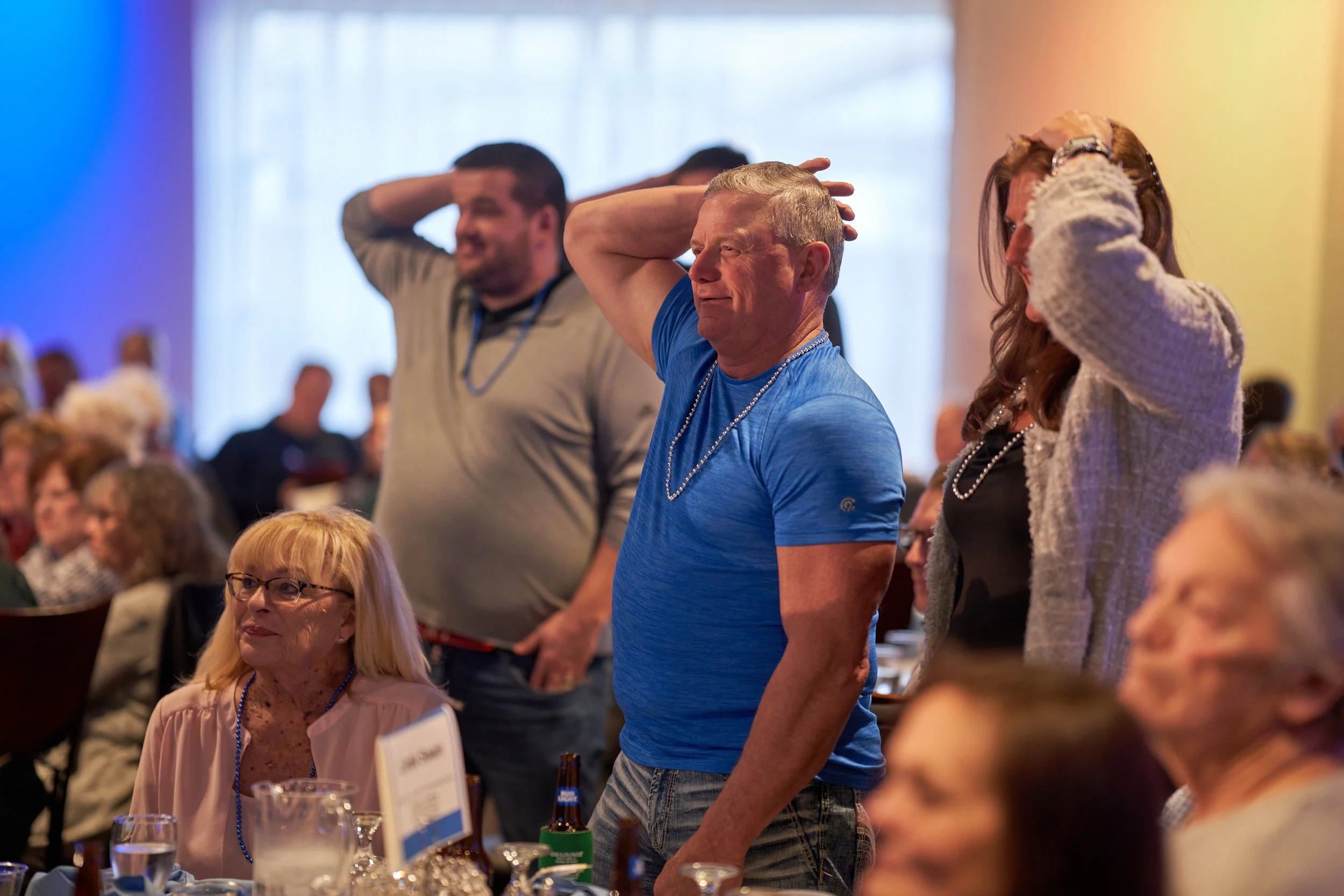 People at an event, some standing with hands on their heads, others seated. Tables with drinks and decorations in foreground.
