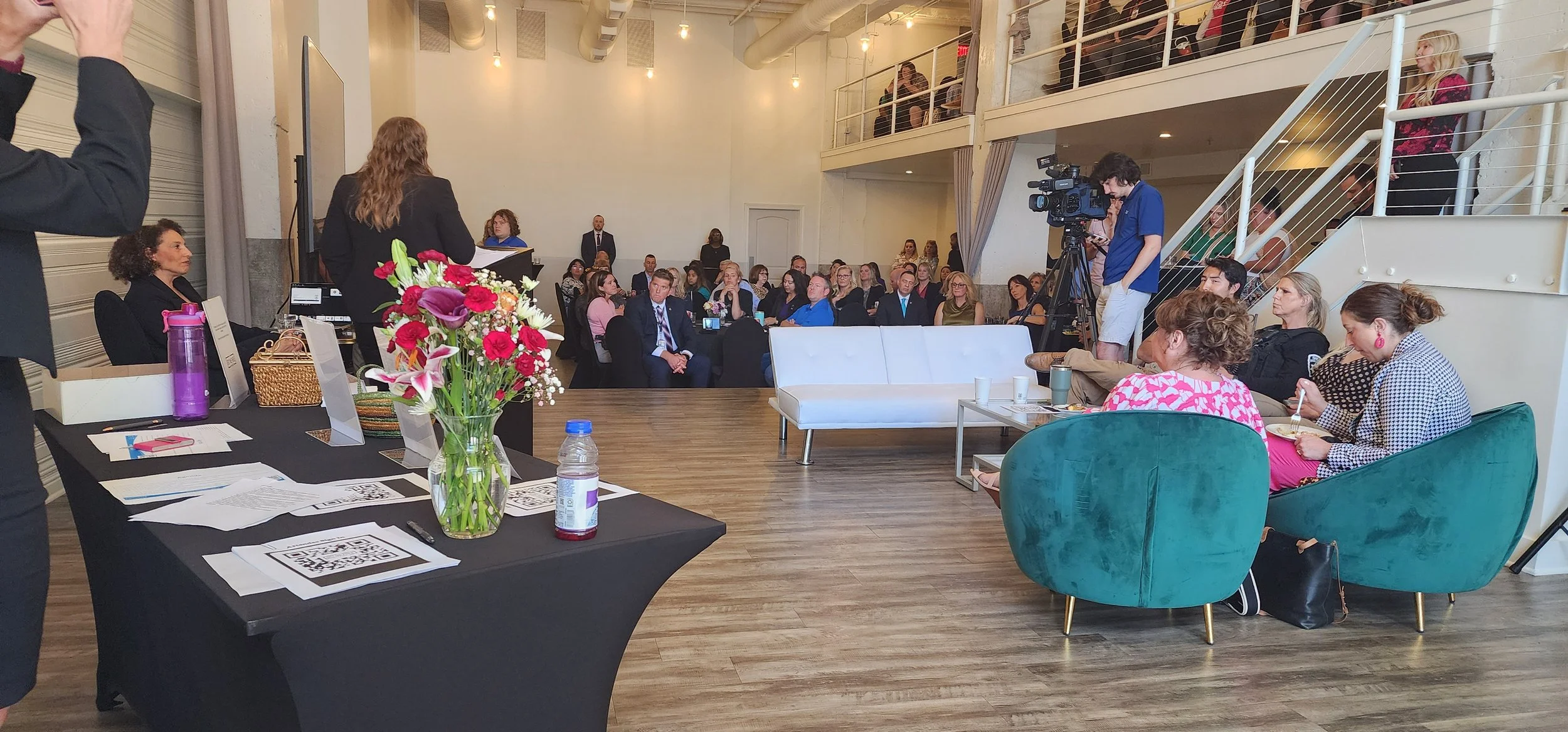A speaker addresses an audience in a stylish venue with modern seating. The room is filled with people sitting and listening attentively. A camera operator is recording the event, and a decorative table with flowers and papers is in the foreground. The room has an open, loft-style design with visible overhead lighting and a balcony.