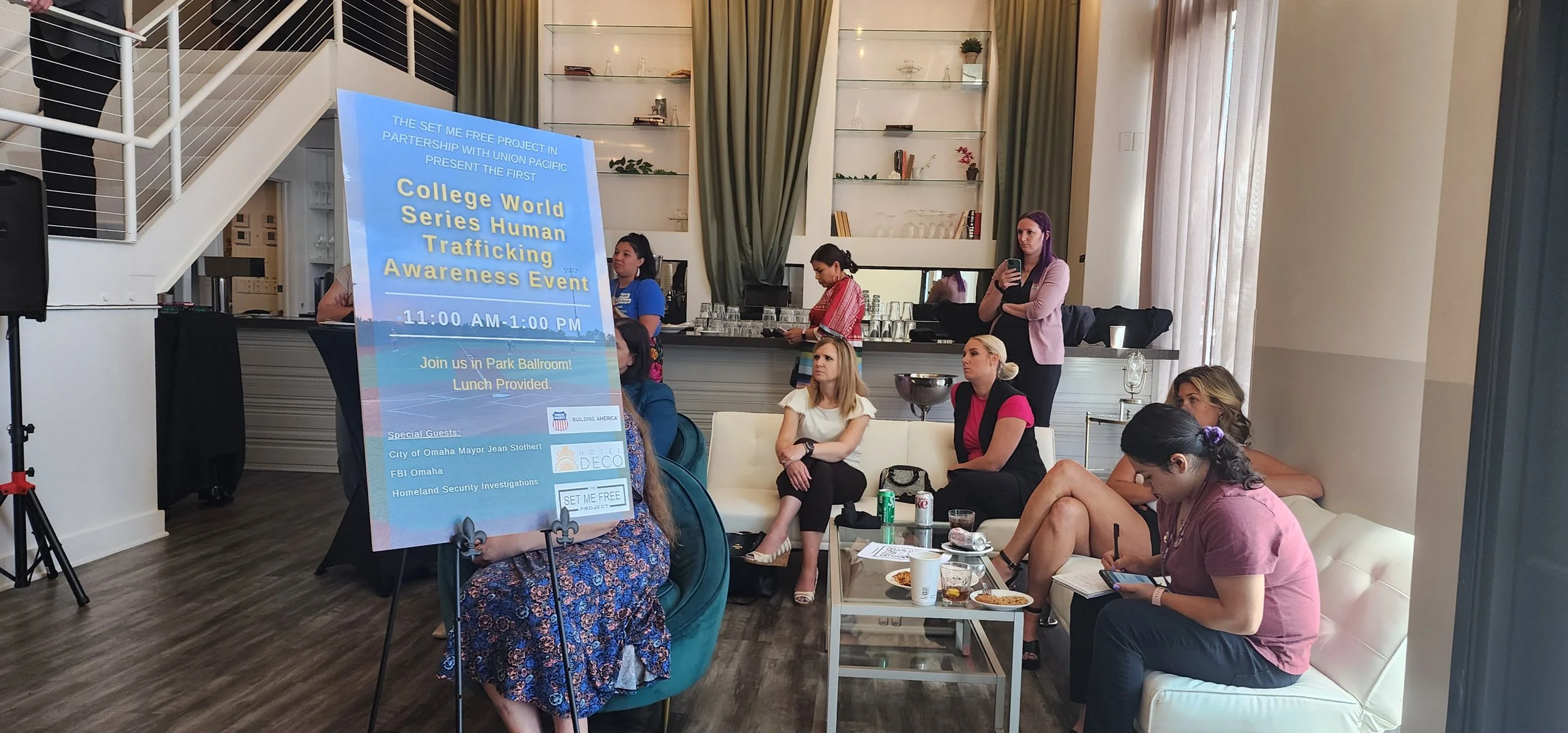 People attending a human trafficking awareness event by "The Set Me Free Project" with a poster in the foreground. The venue includes seating areas and attendees interacting and taking notes. The event features partnerships with Union Pacific and other organizations, hosted at Park Ballroom.