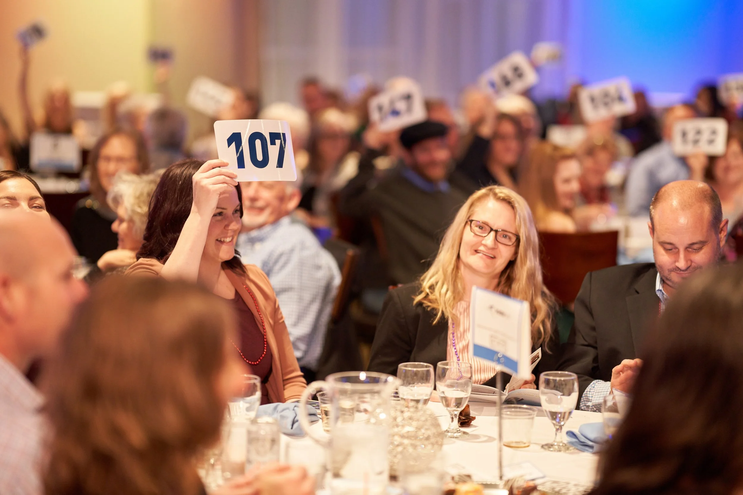 People attending an auction event, sitting at tables, some holding number cards in the air, indicating bids.
